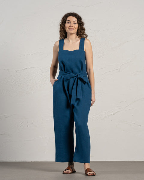 A person with light skin and long, dark curly hair is smiling while standing with their hands in the pockets of a storm blue jumpsuit. The jumpsuit features wide straps and a tie at the waist, and they are wearing brown open-toed sandals.