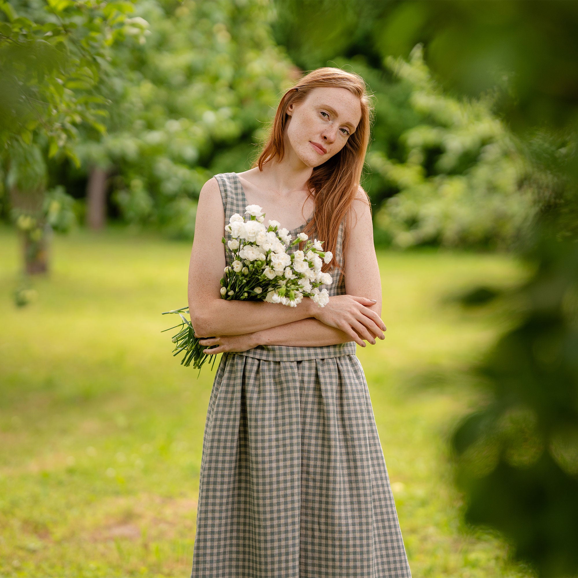 In this picture the model is standing in front of a clothesline with white garments hanging on it. Beside her is a metal bucket full of flowers. She is visible full-height, as well as the full skirt length. The skirt is knee length, matched with a same pattern blouse.