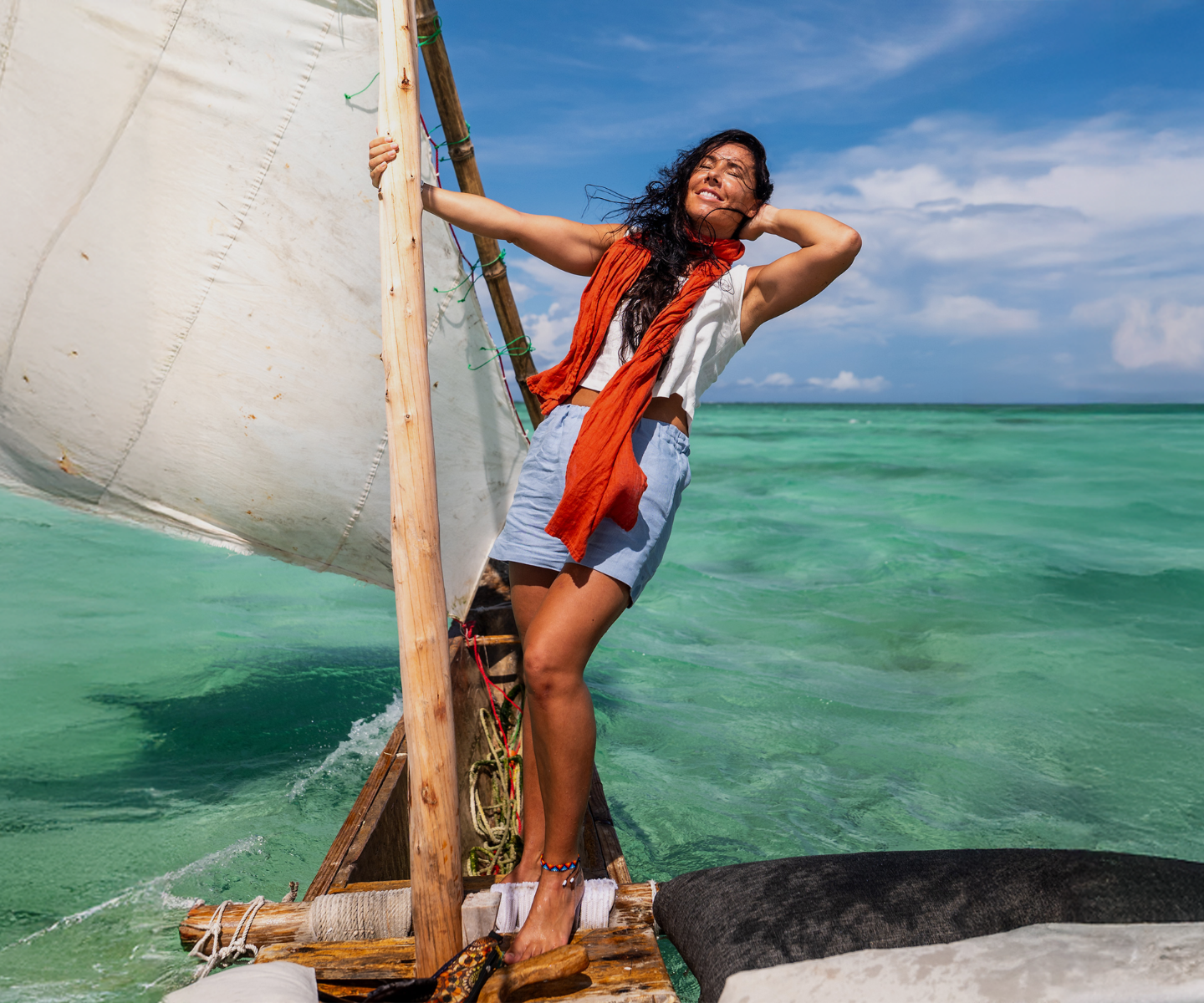A woman standing on a traditional wooden boat, holding onto the mast with a white sail to her side. She is smiling with her eyes closed, enjoying the breeze, with one hand behind her head. She is wearing a white sleeveless top, light blue shorts, and a vibrant orange scarf blowing in the wind. Clear turquoise ocean water and a partly cloudy blue sky fill the background.