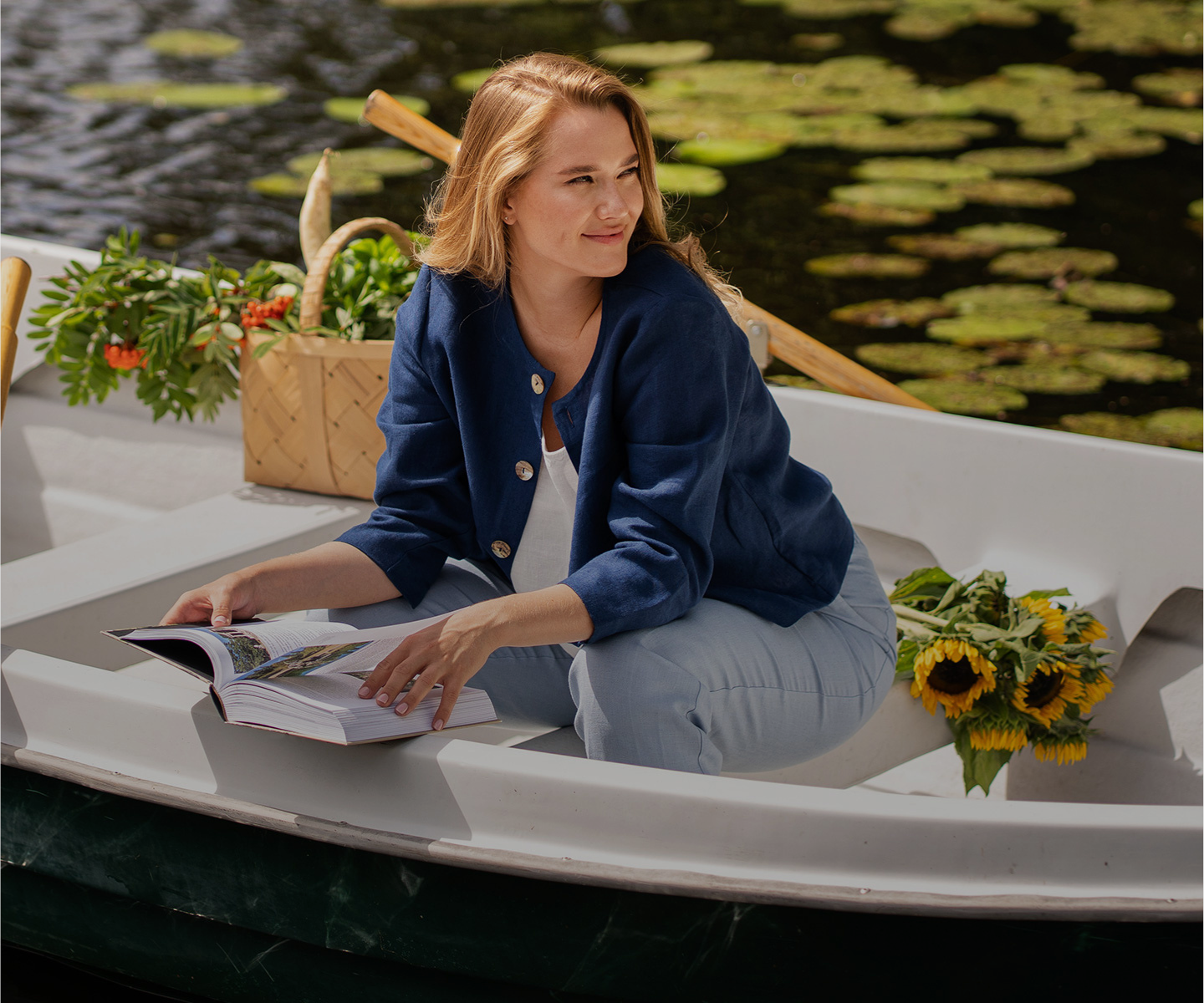 A blonde woman sits in a white rowboat on a lily pad-covered pond, looking thoughtfully off to the right with a gentle smile. She holds an open book in her lap and is wearing a navy blue button-up jacket over a white top with light blue trousers. A wicker basket of greenery and a bouquet of sunflowers rest nearby inside the boat.