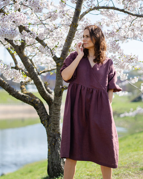 A smiling woman with dark, wavy hair stands outdoors next to a blooming cherry blossom tree. She is wearing a knee-length, shadow purple linen wrap dress with elbow-length sleeves. Her right hand is raised to her face. In the background, there is a body of water and green grass under a bright sky.