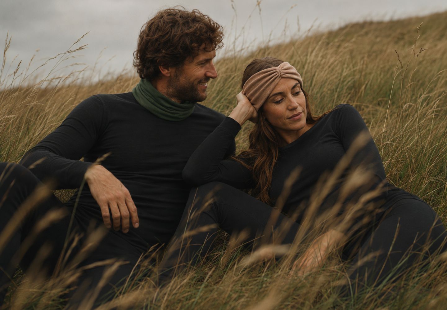 A man and a woman sit comfortably together in a field of tall, dry grass. They are wearing dark, form-fitting long-sleeved clothing and smiling affectionately at one another.