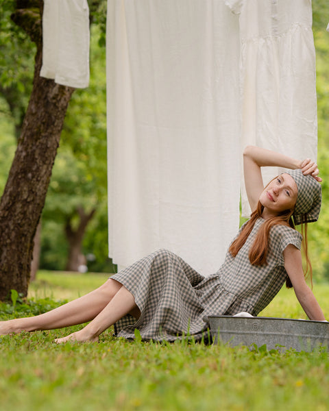 The model is lying in the grass, with her head slightly tilted back and her head lightly touching her forehead. She is wearing a green gingham dress with a matching pattern bandana on her head. She is lying with her legs crossed over each over, near a metal laundry bucket.