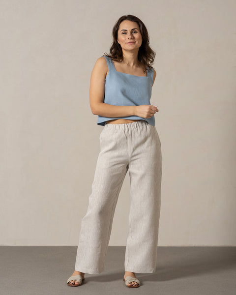 A woman with medium-toned skin and dark, wavy hair stands facing the camera with a slight smile. She is wearing menique linen, sleeveless top with a square neckline and linen wide-legged pants with an elastic waistband. Her hands are casually at her sides, and she is wearing light-colored sandals. She stands on a light gray surface in front of a plain, light beige wall.