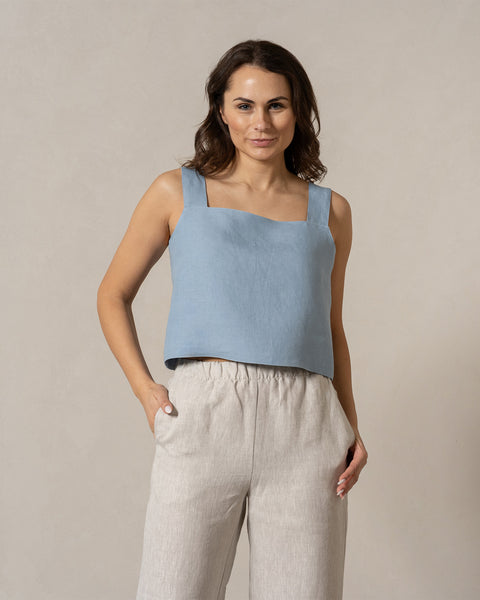 A woman with medium-toned skin and dark, wavy hair looks directly at the camera with a gentle expression. She is wearing a cloudy blue, sleeveless top with a square neckline and natural color, wide-legged pants with an elastic waistband, and her hands are casually placed in her pockets. The background is a plain, light beige color.