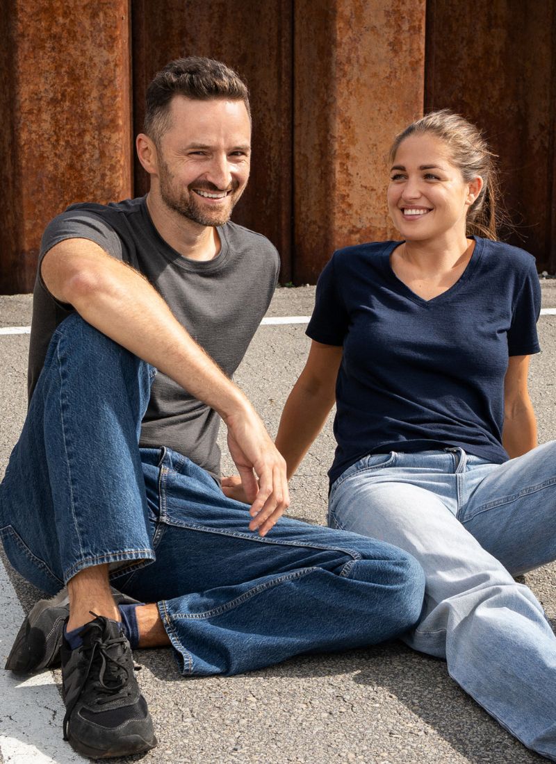 A man and a woman sitting outdoors on a paved surface in front of a rusted metal wall. The man wears a dark gray t-shirt and blue jeans, while the woman wears a navy blue v-neck t-shirt and light-wash jeans.