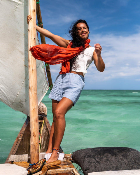 A woman stands on a rustic wooden boat with a white sail against a bright blue sky and turquoise water. She wears a white sleeveless top, light blue shorts, and an orange scarf. The boat has a white cushion and a dark gray cushion.