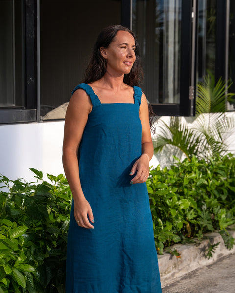 A woman with long, dark hair stands outdoors, wearing linen sundress with a square neckline and thick straps. She is looking off to the side, and lush green foliage surrounds her. The background shows a white building with dark framed windows and more greenery.