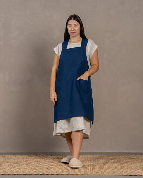 A young woman with dark hair standing in a studio background, with one hand inside the front pocket. She is wearing a linen dress with a linen cross-back japanese apron in storm blue color in size L/XL on top.