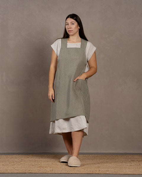 A young woman with dark hair standing in a studio background, with her hand in a front pocket. She is wearing a linen dress with a linen cross-back japanese apron in green check color in size L/XL on top.