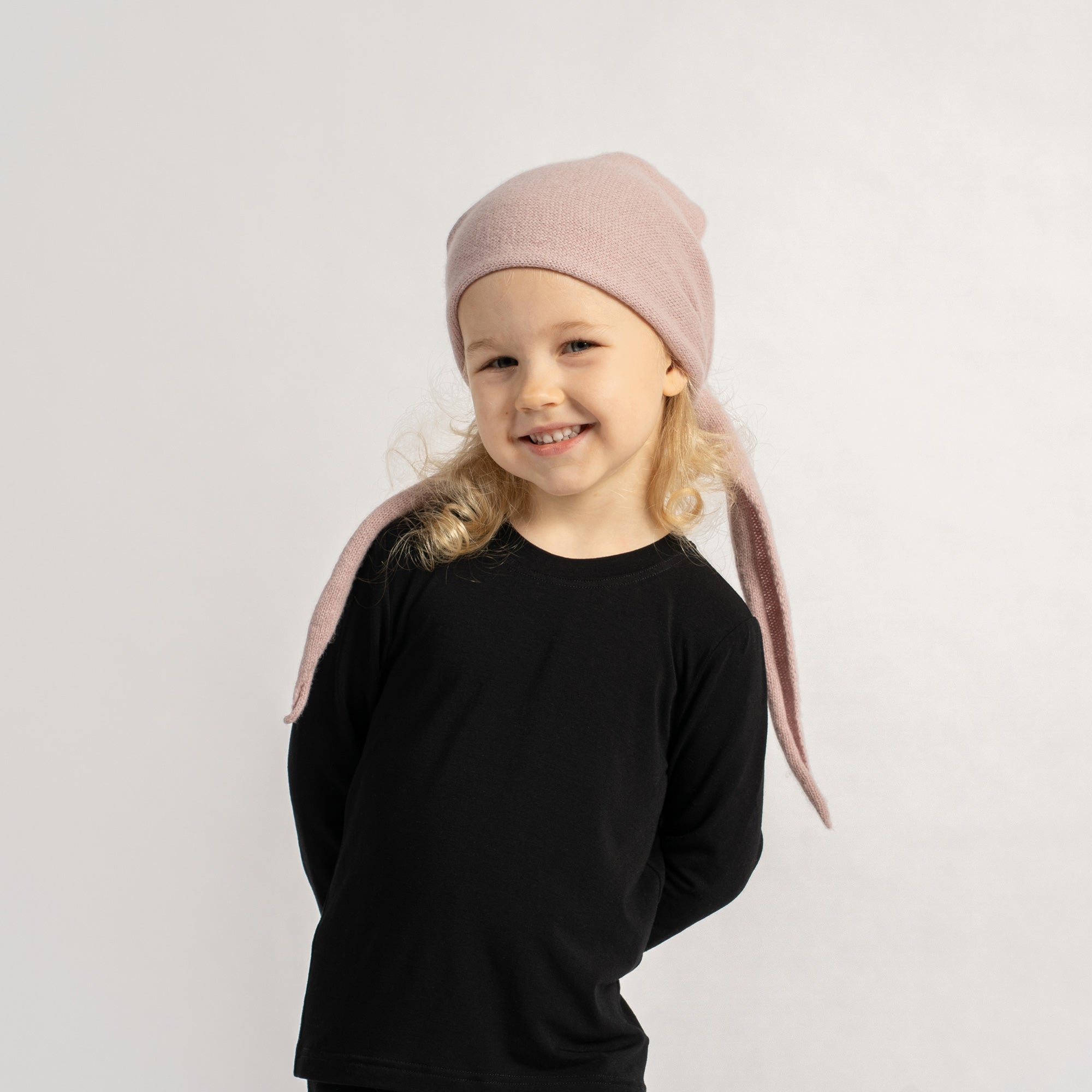 Child wearing a pink knit headscarf, smiling and posing in a black outfit against a plain background.