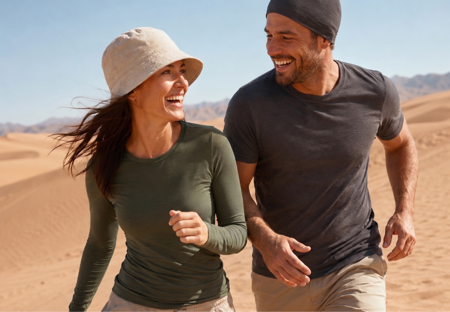 A vibrant lifestyle shot of a happy couple walking together through a sandy desert landscape. The woman is wearing a beige bucket hat, a dark green long-sleeved shirt, and beige bottoms. The man is wearing a grey beanie, a charcoal grey t-shirt, and beige bottoms. Sand dunes and distant mountains form the background under a clear sky.