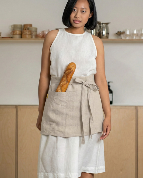 Model wearing a natural linen half apron tied at the waist, baguette tucked into the front pocket, standing in a modern kitchen.