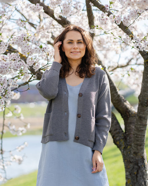 A woman with brown hair, wearing a grey knit cardigan over a light blue dress, stands outdoors next to a tree with white blossoms.