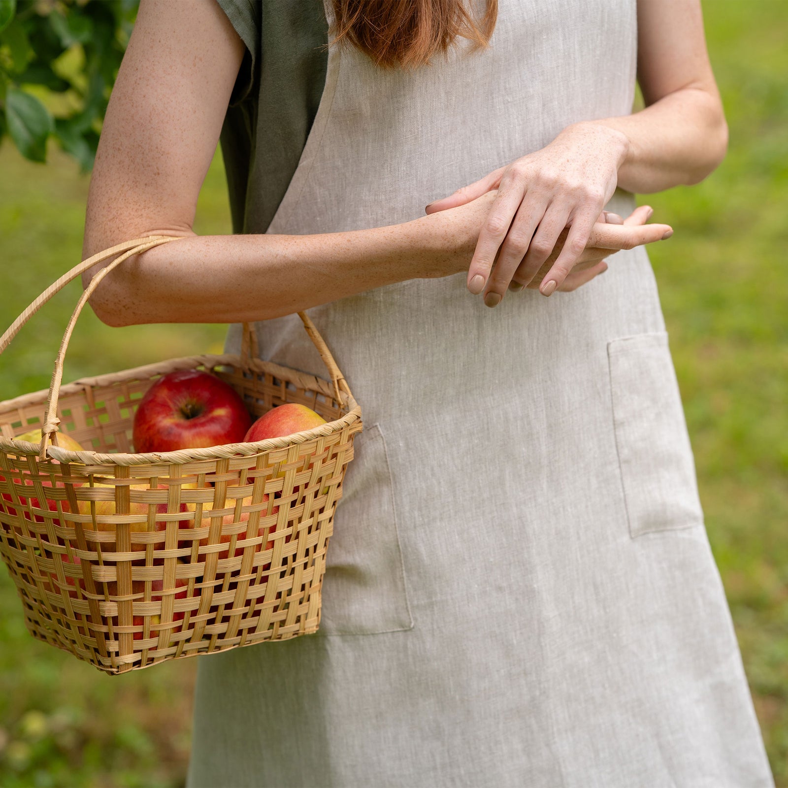 A woman with long red hair wearing a natural color linen apron, standing in a lush green garden and holding a woven basket.