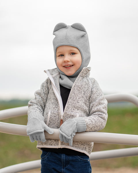 A young child is smiling at the camera while leaning on a light-colored metal structure outdoors. They are wearing a light grey textured zip-up jacket with a white zipper, blue jeans, and matching grey knitted accessories including a balaclava-style hat with two rounded ears on top, and fingerless gloves with a flap that can cover the fingers. The background is a blurred light grey sky.