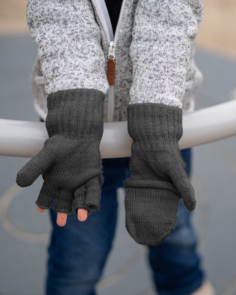 A close-up shot of a person&#39;s hands wearing dark grey convertible mittens. The person is wearing a grey speckled jacket and blue jeans, with their hands resting on a light-colored curved rail. The left glove is in its fingerless form, exposing the fingers, while the right glove has the top flap pulled over, converting it into a full mitten.