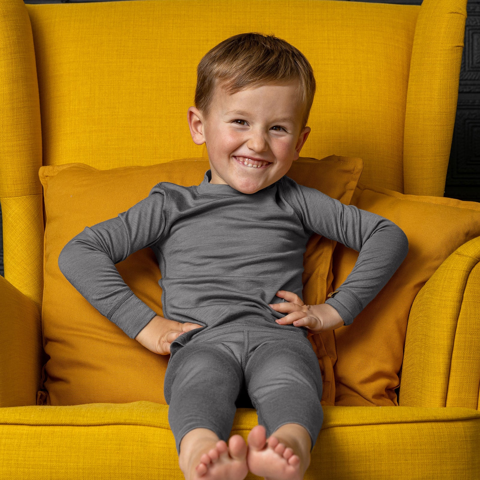 Smiling boy in gray Merino wool base layer set sitting cross-legged on a chair with arms raised.