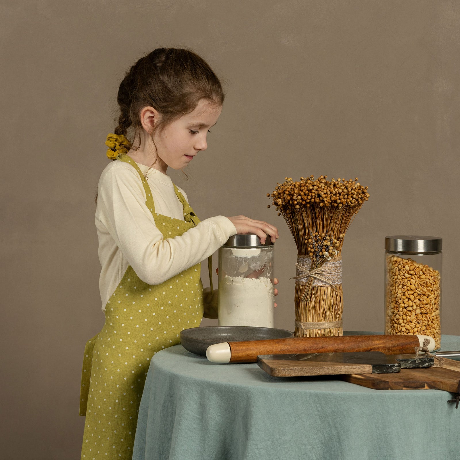 A child wearing a linen apron with leaf dots, standing at a table involved in crafting or baking, with ingredients and baking tools on the table.