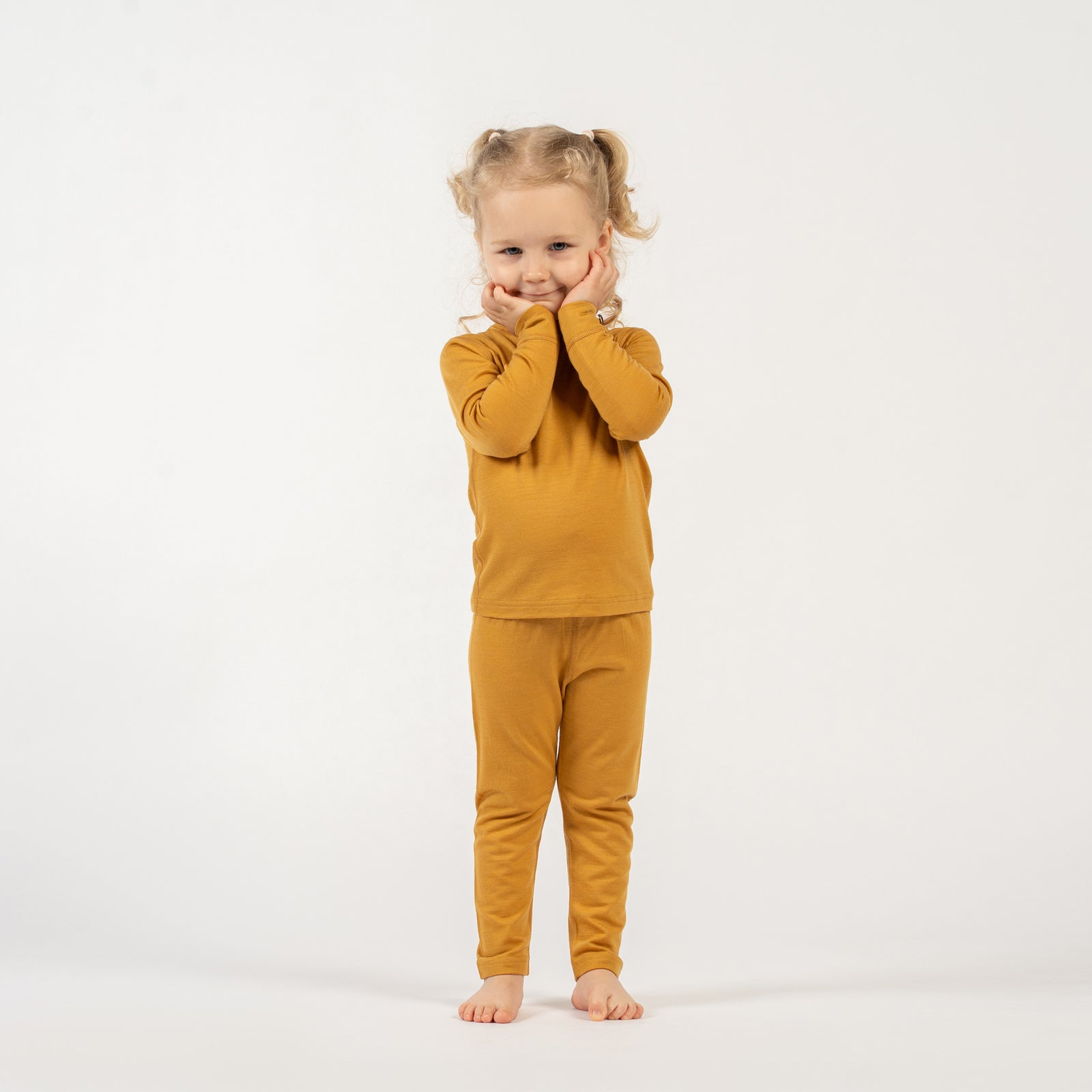 A young child with curly blonde pigtails, wearing a yellow long-sleeved top and matching pants, stands barefoot on a white background, looking directly at the camera with a gentle smile.
