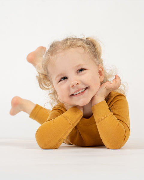 A smiling child with curly blonde pigtails, dressed in a yellow long-sleeved top and matching pants, is lying on their stomach on a white surface. The child&#39;s head is propped up by their hands, and they are looking towards the camera with a joyful expression. Their bare feet are raised slightly in the background.