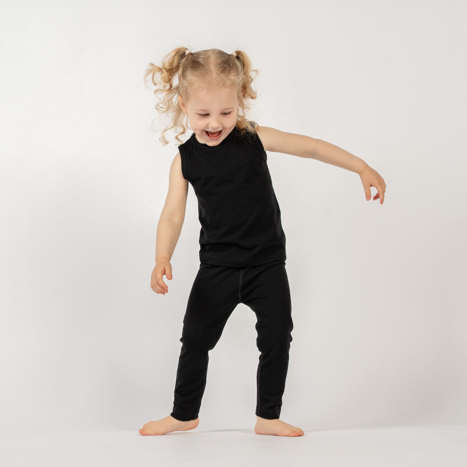 A young child with light curly hair in pigtails is standing barefoot against a plain white background. They are wearing a black sleeveless top and black leggings. The child is looking towards the viewer with an open-mouthed smile, and their arms are outstretched slightly to the sides, as if in mid-movement or about to dance.