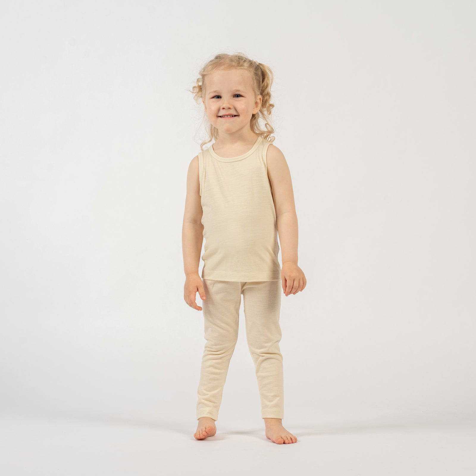 A young child with light, curly hair tied back is standing barefoot against a plain white background. They are wearing a natural colored sleeveless top and matching leggings. The child is smiling widely at the camera with their arms raised above their head, hands clasped together, in a playful pose.