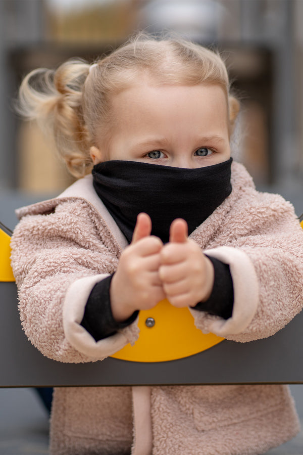 A cheerful child with blonde pigtails looks straight and to the left, peering through a yellow and gray circular opening on a playground. The child is wearing a Black Menique neck gaiter and a fluffy beige jacket, with a slight smile on their face.