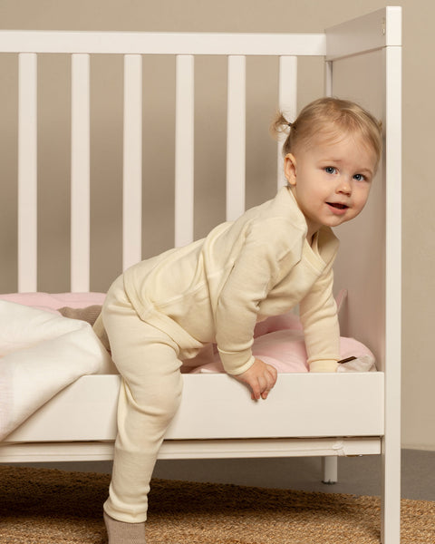 A toddler with blonde hair tied in two small pigtails is standing with one leg inside a white crib and the other leg outside, appearing to climb out. The child is wearing menique long-sleeved top and matching pants, and is looking towards the viewer with a slight smile. Inside the crib, there&#39;s a pink blanket and pillow.