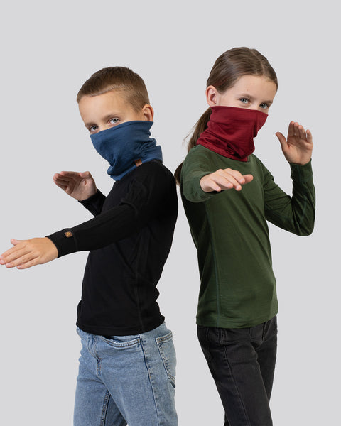 Two kids wearing Merino wool neck gaiters in blue and red, posing playfully in warm, breathable base layers.
