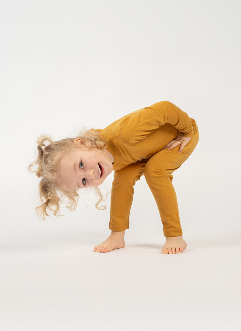A toddler girl with curly blonde hair wearing a mustard yellow long-sleeve one-piece outfit, performing a forward bend on a seamless white studio background.