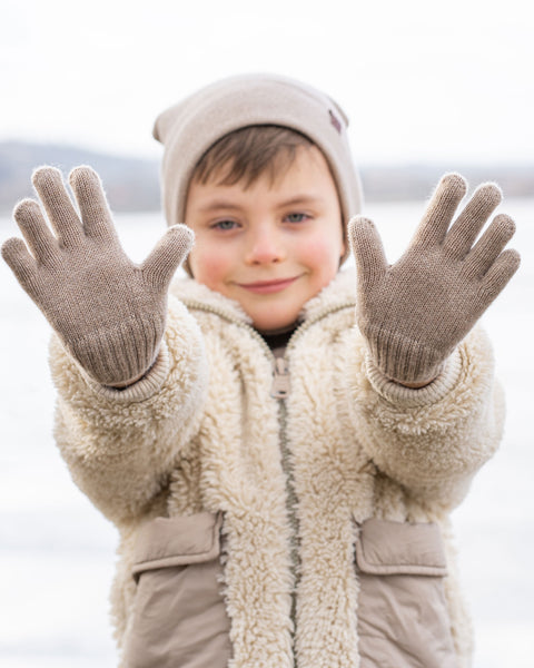 A young child with blue eyes and a faint smile, stands outdoors with both hands outstretched towards the viewer. They are wearing a beige knit beanie with a brown label, a cream-colored sherpa coat with beige pockets, and light brown knitted gloves. The background is a blurred, bright, wintry landscape.