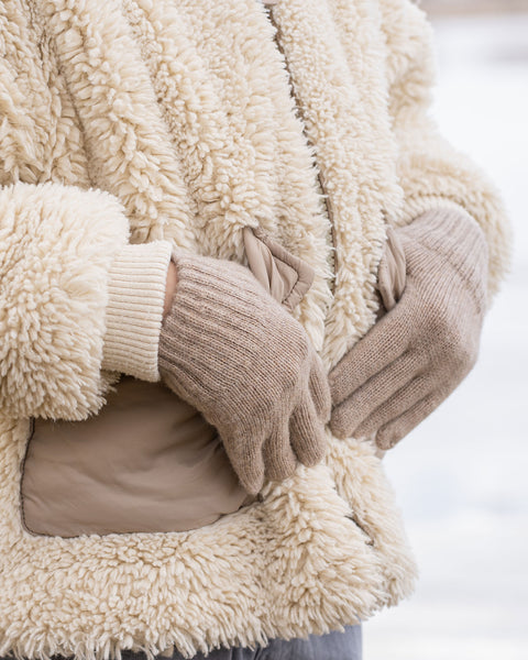 A person wearing a cream-colored sherpa jacket with beige pockets and a light-colored ribbed cuff is adjusting their light brown knitted gloves. The background is blurred and bright, suggesting an outdoor winter setting.
