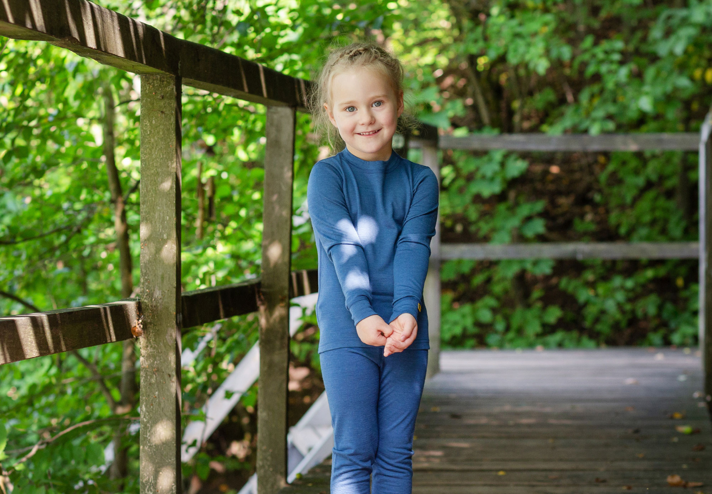 A smiling young girl wearing a blue merino wool base layer set from menique stands on a wooden bridge surrounded by lush green trees and dappled sunlight.