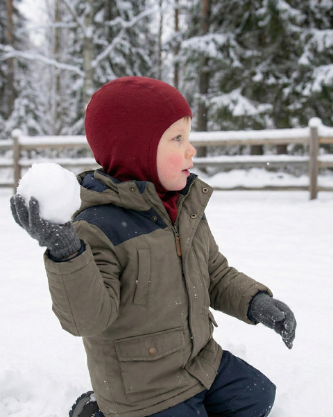 Young child wearing a warm winter jacket and a burgundy merino wool balaclava, holding a large snowball while kneeling in fresh snow in a forested winter landscape.