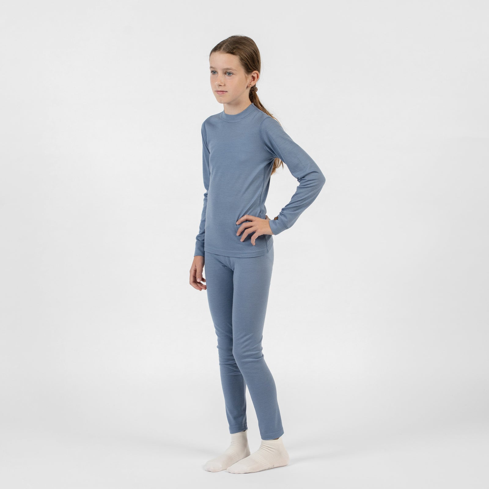 A close-up side view of a child's lower half wearing fitted dusty blue merino bottoms and cream-colored socks, with hands resting near the waistband.