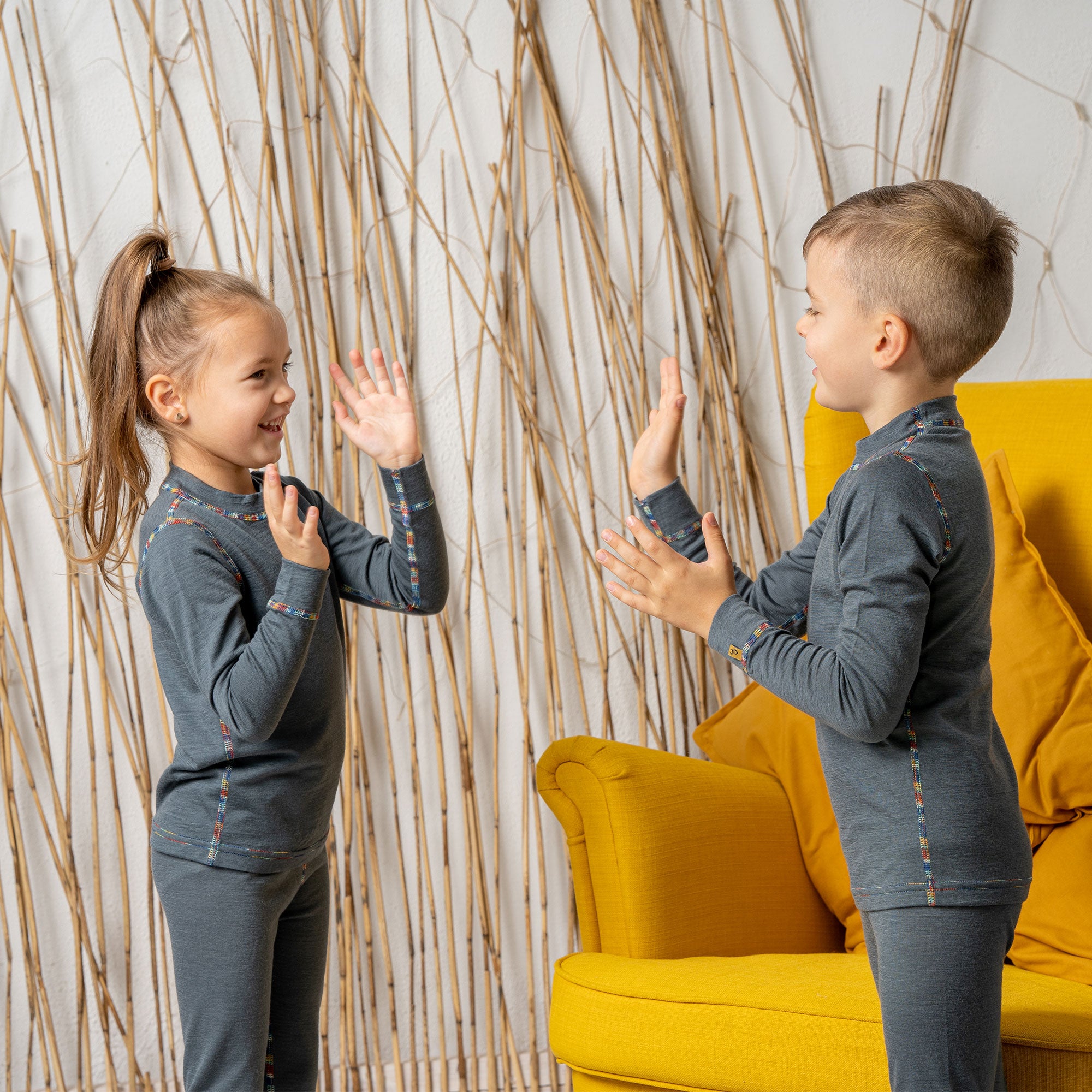 A boy sitting in a yellow armchair, wearing the menique Kids' 160 Merino Pants in Perfect Grey color. He has his hands raised above his head, being cheerful.