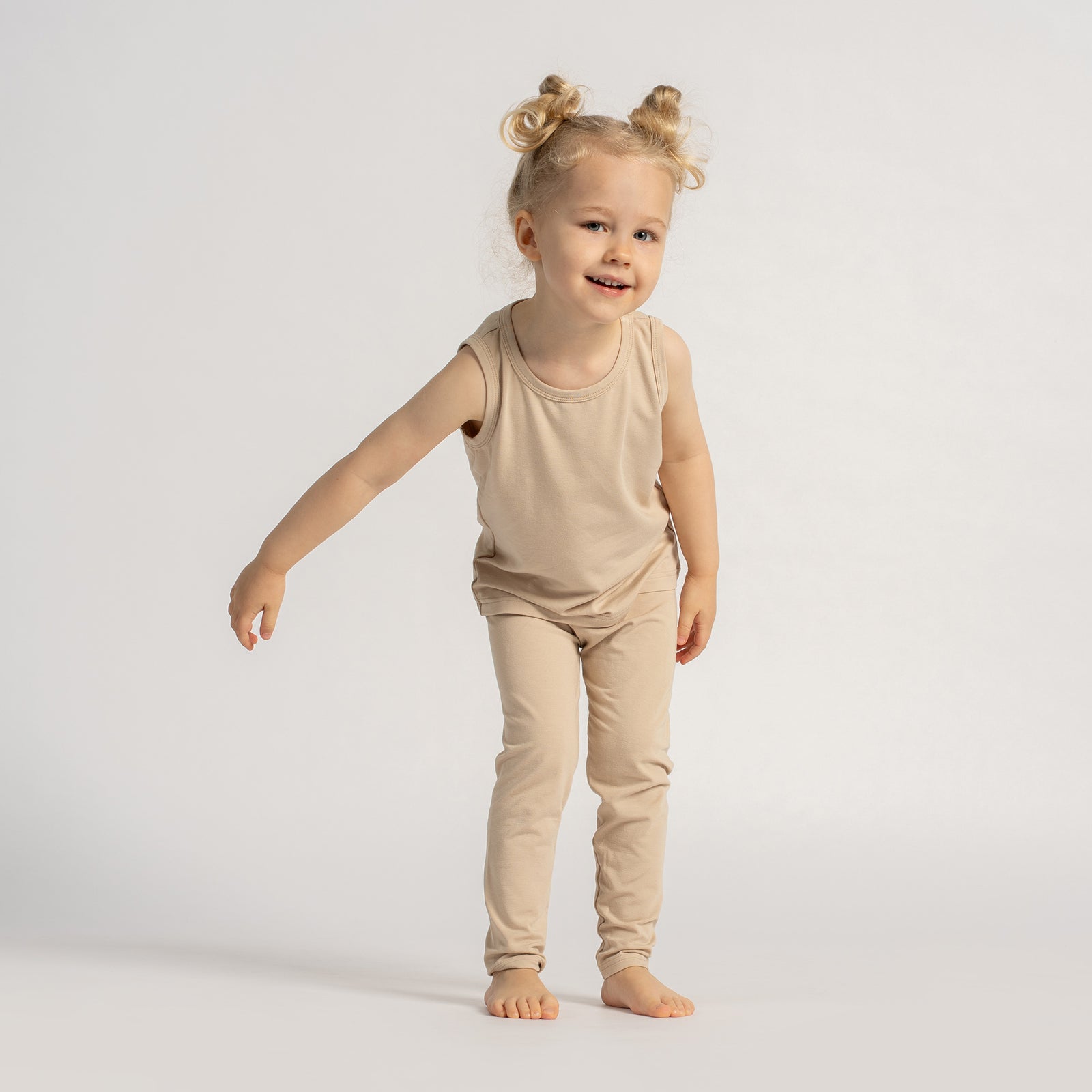 Smiling toddler wearing a beige sleeveless top and leggings set, standing barefoot and playing in front of a light background.