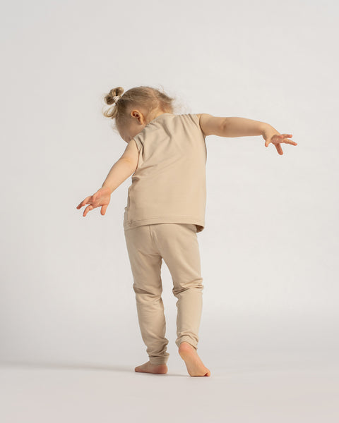 Little girl in a beige sleeveless top and leggings set seen from the back, walking barefoot on a light background.