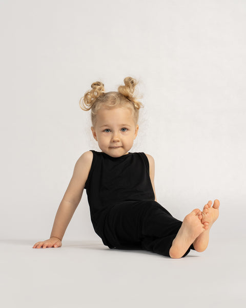 Toddler sitting on the floor wearing a black sleeveless top and leggings, looking forward with a calm expression.