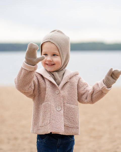 A young child is standing on a sandy or dirt beach with a wide smile, looking towards the right side of the frame with a joyful expression. They are wearing a light brown balaclava-style hat, a light pink or beige textured jacket with large buttons and a collar, dark blue jeans, and matching light brown mittens. Their arms are raised slightly, with hands open. In the background, there&#39;s a body of water under a light, possibly overcast, sky.