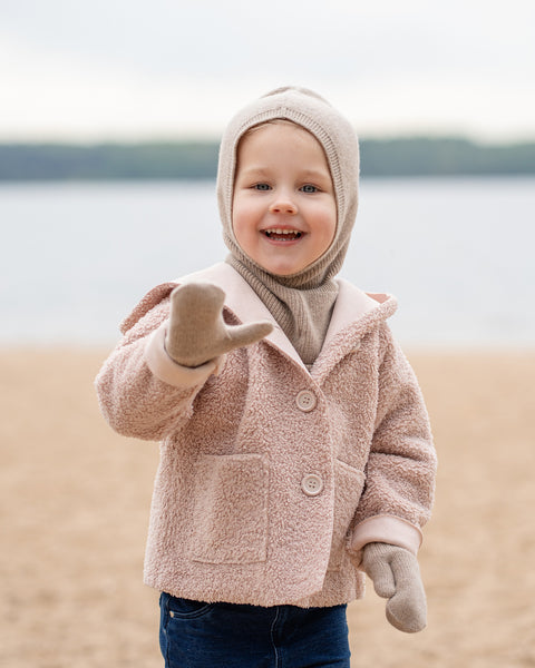 A young child is standing on a sandy beach and looking directly at the camera with a wide, happy smile. They are dressed in a light brown balaclava-style hat, a light pink or beige textured jacket with large buttons and a collar, dark blue jeans, and matching light brown mittens. Their right arm is raised slightly, with the hand in an open gesture. In the background, there is a calm body of water and a faint shoreline on the opposite side, under an overcast sky.