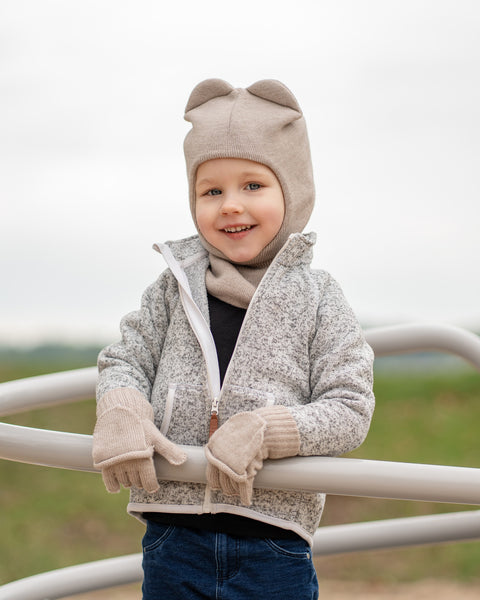 A young child with blue eyes and a wide smile, is leaning on a metal railing outdoors. They are wearing a beige balaclava with ears, a matching scarf, beige gloves, a grey speckled zip-up hoodie, and blue jeans, looking directly at the camera with a blurred green and grey background.