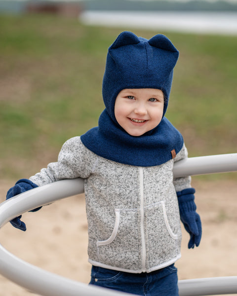 A young child with blue eyes and a slight smile, is leaning on a metal railing outdoors. They are wearing a dark blue balaclava with ears, dark blue gloves, and a grey speckled zip-up hoodie. The background is a blurred outdoor scene with green grass and water.