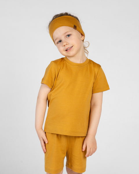 A young child standing against a plain light grey background, smiling with their head tilted slightly to the side. The child is wearing a matching set of mustard yellow clothing, including a short-sleeve t-shirt, shorts, and a wide headband.