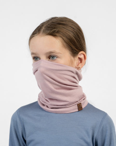 A close-up side view of a young girl wearing a dusty pink merino long neck gaiter pulled over her nose and mouth, paired with a dusty blue long-sleeve shirt.