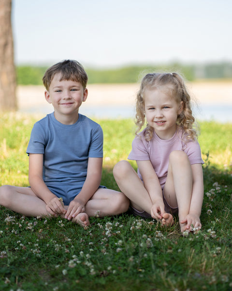 A young boy and girl sit together on a patch of grass with clover. The boy, on the left, sits cross-legged in a blue t-shirt and shorts set. The girl, on the right, sits with her knees bent, wearing a light pink t-shirt and shorts set. Both are barefoot and smiling at the camera, with a body of water faintly visible in the background.
