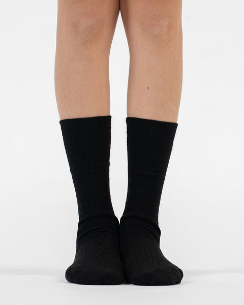 A front view of lower legs standing flat, modeling black ribbed merino crew socks.