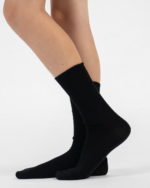 A side-profile view of a person standing, wearing black ribbed merino crew socks, with the back foot slightly lifted.