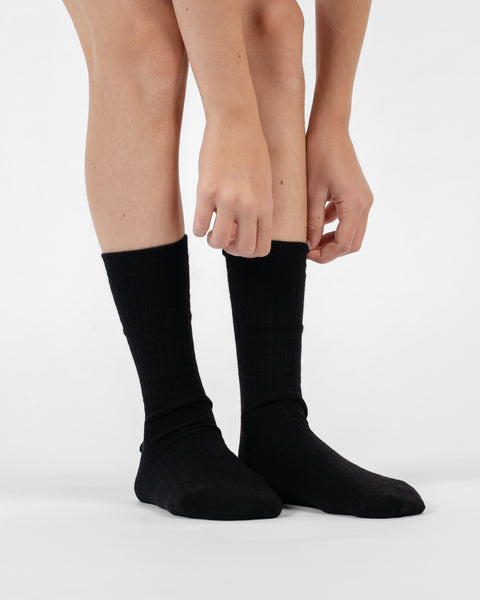 A front-facing view showing hands adjusting the top cuff of black ribbed merino crew socks on a person's legs.